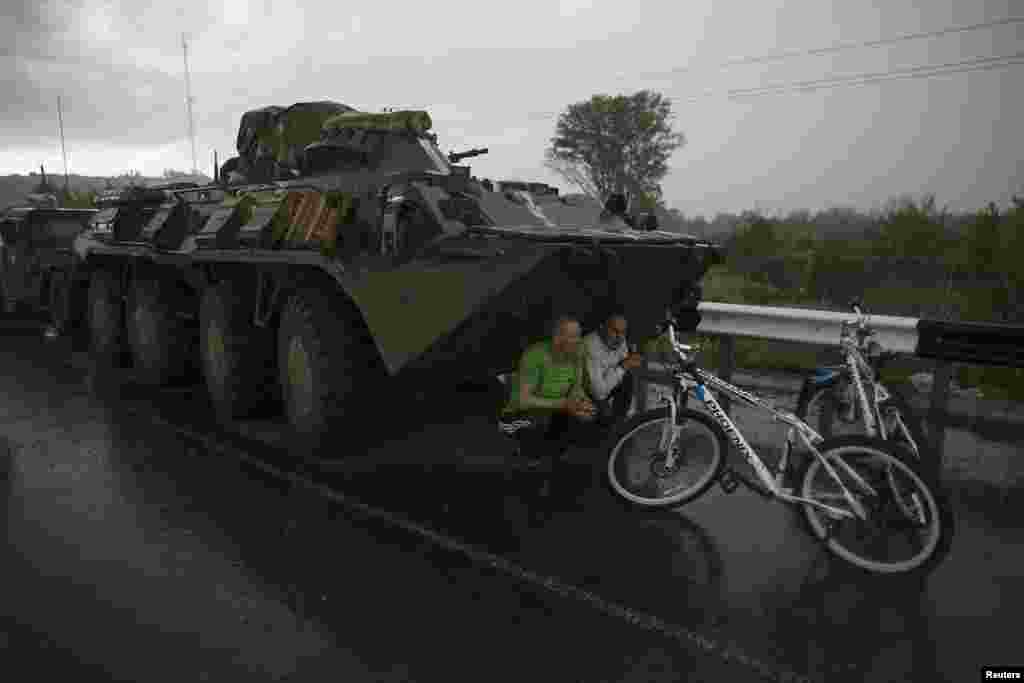 Dua warga sipil menemukan tempat untuk berlindung dari hujan di bawah kendaraan lapis baja tentara Ukraina di sebuah pos pemeriksaan dekat kota Slaviansk. 