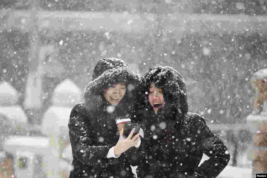 Dua perempuan Korea tersenyum sambil melihat foto dari ponsel mereka saat hujan salju di Seoul, Korea Selatan.