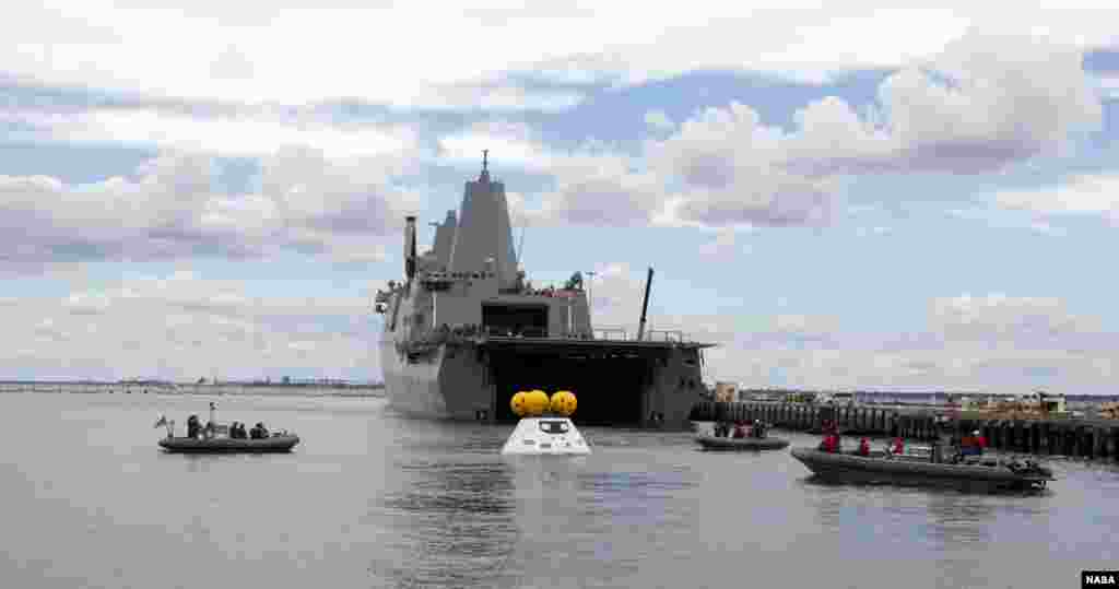 With the U.S. Navy's well deck ship USS Arlington stationed against its pier at Naval Station Norfolk in Virginia, divers in small boats approach a test version of NASA's Orion crew module during the stationary recovery test. The test is helping to ensure that when Orion returns from deep space missions and splashes down in the Pacific Ocean.
