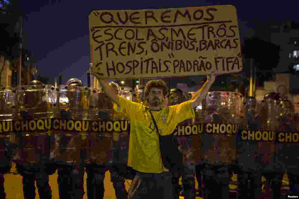 A protester holds a sign that reads "We want schools, subways, trains, buses, boats, hospitals of FIFA standard" in front of riot police during a demonstration against the public spending for the 2014 World Cup, in Rio de Janeiro, June 15, 2014.