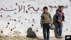 Un niño sirio sostiene una pistola de juguete mientras juega al fútbol con otros entre edificios destruidos en lo que un graffiti dice "Siria al-Assad" en la ciudad vieja de Homs, Siria, el viernes, 26 de febrero de 2016.