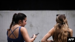 Dua remaja perempuan duduk di kursi sambil menatap telepon genggam mereka di pusat Kota Sydney, Australia, pada 7 November 2024. (Foto: AFP/David Gray)
