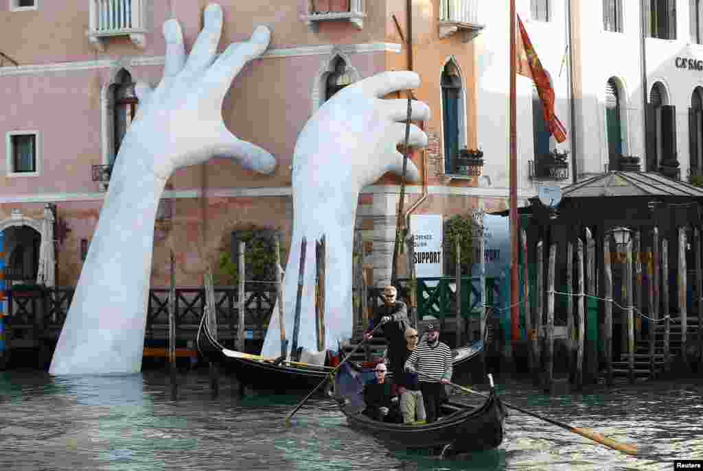 Para turis menaiki gondola di dekat karya seni karya seniman Italia Lorenzo Quinn di istana Ca' Sagredo, Venesia, Italia.