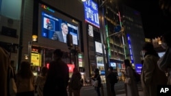 Japanese Prime Minister Yoshihide Suga, on a screen, speaks at a news conference in a live broadcasting shown in a big screen on a building as people walk across an intersection in the Shinjuku district of Tokyo on March 18, 2021.